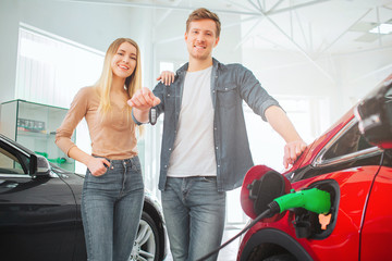 Young smiling family buying first electric car in the showroom. Attractive man holding car key while standing between his wife and eco-friendly car. Electric car sale concept