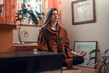 Young woman meditating, exercising yoga in a lotus position at her home, sitting next to a window