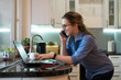 © Vagengeim - Business woman in kitchen at home. She is talking on cell phone and looking at laptop screen. Girl freelancer makes plan for day. Good morning. Self-employed person