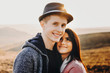 © Strelciuc - Portrait of cute adult caucasian couple where man is wearing a hat and looking at camera smiling while embracing with a hand his girlfriend with is looking at him against sunset while dating.