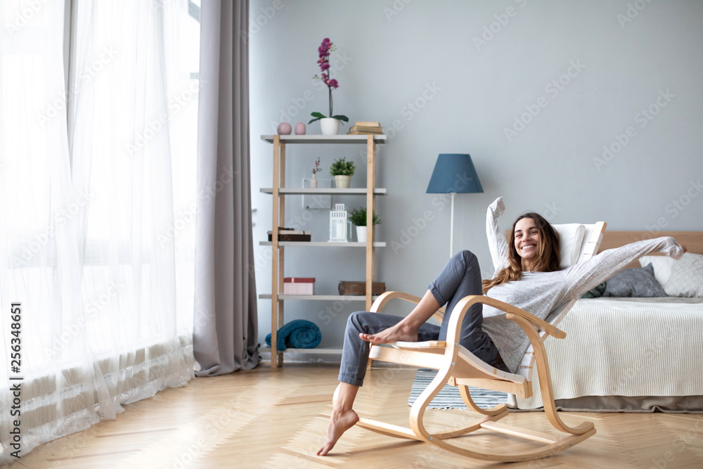 Happy woman resting comfortably sitting on modern chair in the living room at home. - obrazy, fototapety, plakaty 