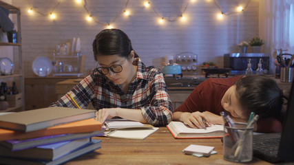 front view of tired student studying with laptop computer at home in midnight fell asleep on wooden table. smart hard working female asian classmate still preparing exam writing notebook in kitchen