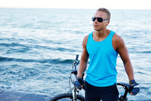 Portrait Of A Handsome Blond Athletic Man With Bicycle Outdoors On The Beach. Strong Sports Guy In Sunglasses Posing Against The Sea. Summer Beach Sports Vacation.