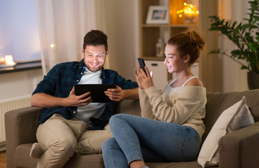 leisure, technology and internet addiction concept - happy couple with tablet computer and smartphone at home in evening