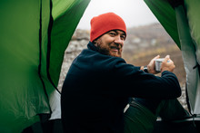 Portrait Of A View From Tent Entrance Of Young Traveler Man Smiling And Relaxing In Mountains Near To Bonfire. Rear View Of Hiker Male Sitting In The Camping Tent, Drinking Hot Beverage. Travel People