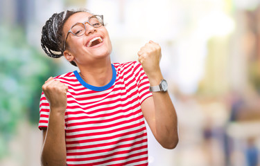 Sticker - Young braided hair african american girl wearing glasses over isolated background celebrating surprised and amazed for success with arms raised and open eyes. Winner concept.