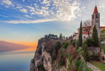 Wall Mural - View of the church (Chiesa di San Giovanni Battista) of the city Tremosine (Tremosine sul Garda) and Lake Garda in the rays of the setting sun. Winter period. Lombardy, Italy