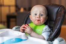 A Funny Adorable Open-eyed Baby Boy Of 10 Months Eats Strawberry Sitting In The Baby Chair At Home In The Evening Wearing Feeding Bib Is Surprised By The Taste Of The First Baby Food 