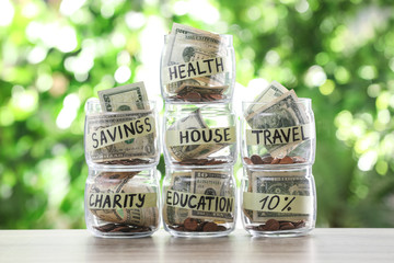 Glass jars with money for different needs on table against blurred background