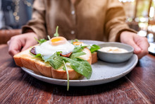 A Woman Holding A Plate Of Breakfast Sandwich With Eggs, Bacon And Sour Cream On Wooden Table