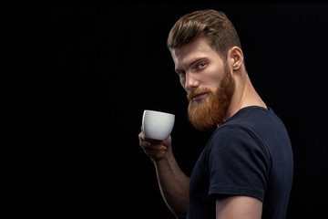 Wall Mural - Beautiful young bearded man drinking coffee and looking at camera Resting handsome perfect hairstyle man drinking espresso coffee. Man holding cup of coffee in hand over black background.