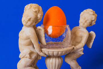 Wall Mural - A statue of angels on a blue background holding a glass, cracked glass, inside an Easter egg orange.  The concept of tradition of Easter.