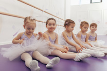 Group Of Cute Little Girls Wearing Ballet Tutus Sitting On The Floor At Dance Studio. Adorable Little Ballerinas Rest After Ballet Class. Health, Gymnastics, Activity Concept
