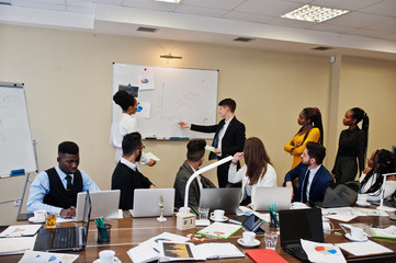 Mixed race business coach presenting report standing near whiteboard pointing on sales statistic shown on diagram and chart teach diverse company members gathered together in conference room.