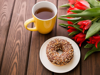 donut on white plate with red tulips and cup of tea on a brown wooden background. Breakfast in summer time, top view, copy space