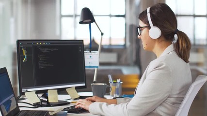 business, programming and technology concept - female programmer in headphones working with script on computer screen and listening to music at office