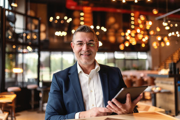 Middle aged man dressed smart casual holding tablet and looking at camera while sitting in cafeteria.