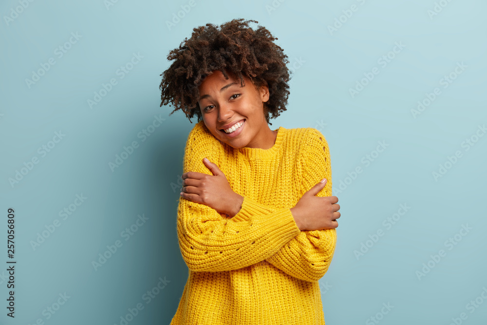 Headshot of pleased dark skinned woman hugs herself, smiles from pleasure, likes new yellow sweater she bought, has Afro hairstyle, crosses hands, recalls romantic moment, isolated on blue wall - obrazy, fototapety, plakaty 