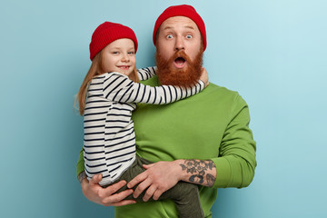 Wall Mural - Shocked fashionable dad stares at something unbelievable, has thick ginger beard, carries small daughter who embraces him with love, play game together, have spare time, isolated over blue background