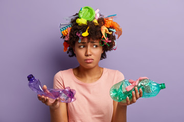 Sad bothered woman holds two crumpled plastic bottles, feels dejected, cleans up earth from contamination, dressed in casual clothes, has litter left in hair, stands over purple studio wall. Reuse