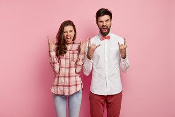 Wall Mural - Photo of punky joyful man and woman hear music of favourite band, gesture actively, show rock n roll gesture, being fan of particular genre, come on party, wear stylish clothes, isolated on pink wall