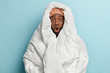 Photo of stupefied dark skinned man wrapped in white blanket, feels cold in room, cant get asleep, stands over blue background. Morning awakening concept. Unhappy surprised adult guy under sheet