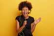 © WHstudio Leushin N - Amused black young woman with curly hair, shakes hands, enjoys cool music, holds smart phone, perfect sound in headphones, has heartbreaking moment, listens favourite track, isolated on yellow