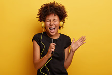Amused Black Young Woman With Curly Hair, Shakes Hands, Enjoys Cool Music, Holds Smart Phone, Perfect Sound In Headphones, Has Heartbreaking Moment, Listens Favourite Track, Isolated On Yellow