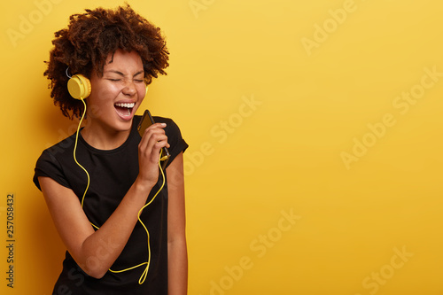 Delighted happy dark skinned emotional woman feels herself as real star, dances and sings along music, uses smart phone as microphone, modern headphones, stands over yellow studio wall, enjoys life
