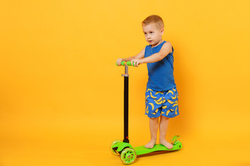 Kid boy 3-4 years old wearing blue beach summer clothes on scooter isolated on bright yellow orange wall background, children studio portrait. People, childhood lifestyle concept. Mock up copy space.