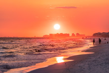 Couple Walking On Shore With Dramatic Orange Red Pink Sunset In Santa Rosa Beach, Florida With Pensacola Coastline Coast Cityscape Skyline In Panhandle With Ocean Gulf Mexico Waves