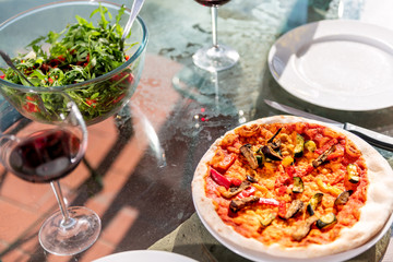 Fresh whole pizza and salad on glass table terrace outside in Italy with tomato sauce vegetables and two glasses of red wine