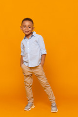 Wall Mural - Vertical studio shot of attractive little boy of African origin posing isolated against blank yellow wall background, wearing stylish clothes, smiling confidently, keeping hands in pockets of jeans