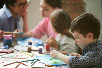 Wall Mural - Side view portrait of cute little boy painting pictures in art class with group of children, copy space