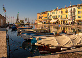 Wall Mural - Scenic view of the Old Port of the medieval town of Lazise on the shore of Lake Garda with the typical colored houses and moored boats in a sunny day, Veneto, Italy