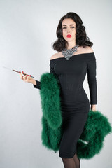 Wall Mural - charming graceful pin up girl in a black evening dress and a green fluffy boa in her hands stands and smokes a cigarette with a mouthpiece on a white solid Studio background