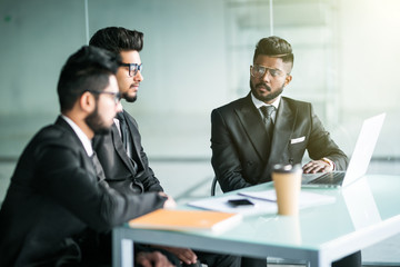 Three indian businessmen sitting around table meeting in modern open plan office