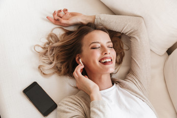 Poster - Pretty blonde woman posing sitting indoors at home listening music with earphones.