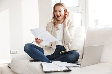 Poster - Beautiful blonde woman posing sitting indoors at home using laptop computer talking by mobile phone.