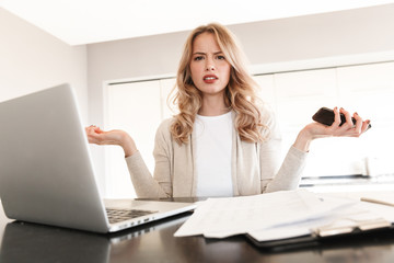 Wall Mural - Confused blonde woman posing sitting indoors at home using laptop computer holding mobile phone.