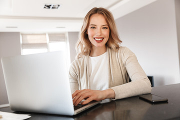 Wall Mural - Beautiful blonde woman posing sitting indoors at home using laptop computer.