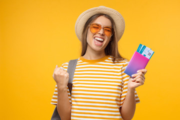 Summer shot of young happy female traveller holding passport with flight tickets screaming if she is a winner, isolated on yellow background