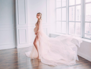 cute young slim lady with tummy in spacious room with white walls and large windows, pregnant girl with blond hair twisted in a long pink gentle lace peignoir with flying train posing for camera