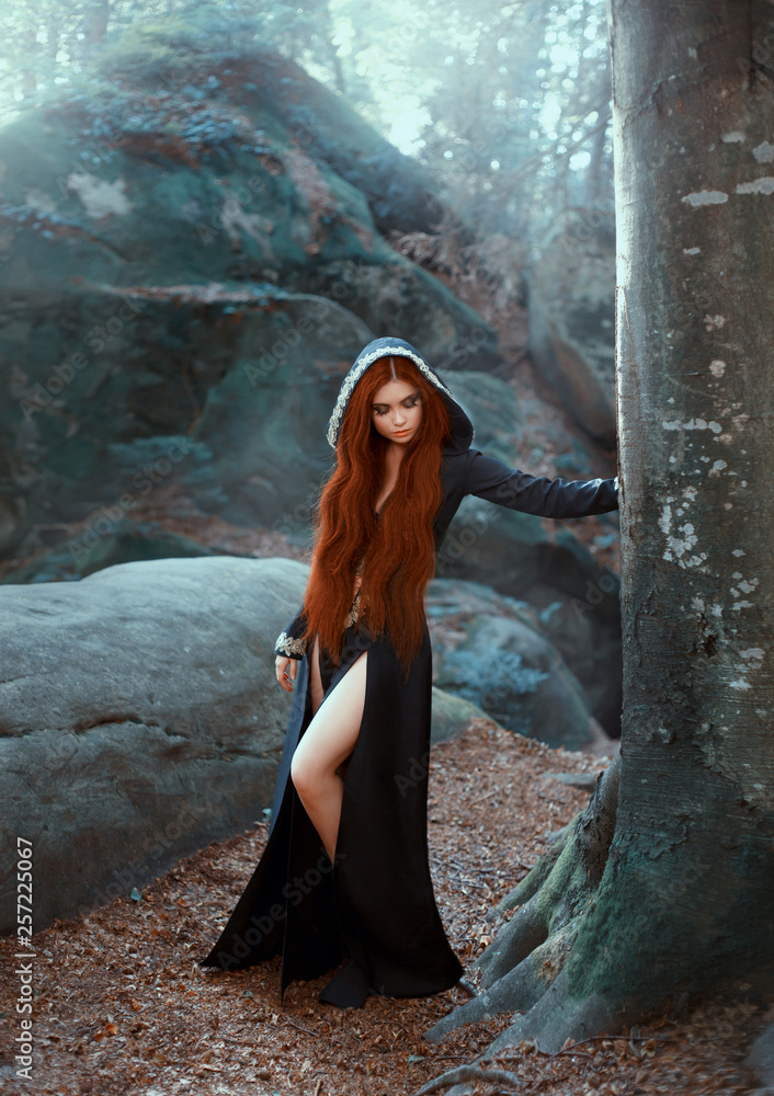 Photo Stock mysterious girl in long black dress with cut for bare legs and  hood stands by tree, dark priestess with bright red hair listens to voices  of frosty forest, winter ice