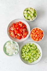 Wall Mural - Fresh chopped vegetables in ceramic bowls on a kitchen white table for preparing vegetarian meal. Top view, copy space.