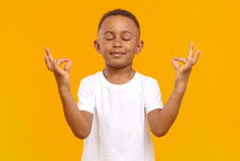 Peaceful Calm Little Boy Of African Appearance Wearing Casual T-shirt Keeping His Eyes Closed And Smiling, Meditating Isolated In Studio. Conscious Dark Skinned Schoolboy Practicing Meditation