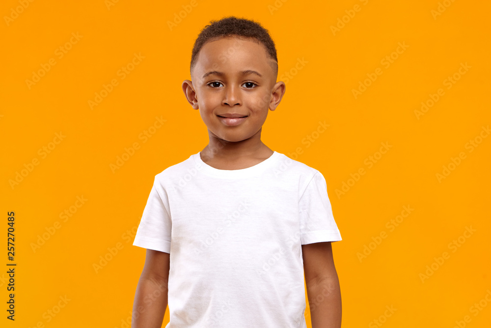 Isolated image of cute adorable dark skinned schoolboy wearing white t-shirt posing in blank yellow studio with confident cheerful smile because he received high marks for maths test at school - obrazy, fototapety, plakaty 