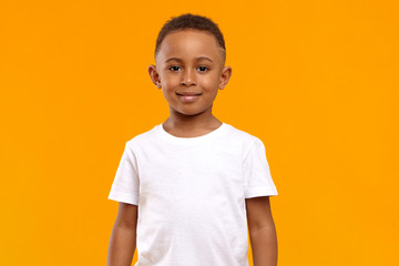 isolated image of cute adorable dark skinned schoolboy wearing white t-shirt posing in blank yellow 