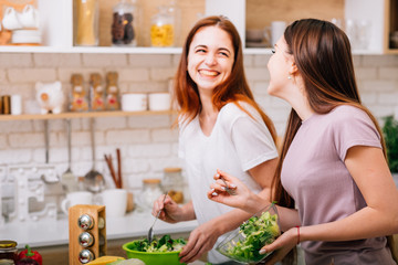 Dieting together. Female cooking fun. Two young women with salad bowls laughing loud.