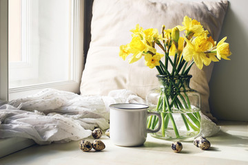 Cozy Easter, spring still life scene. Mug of coffee, wooden plate, quail eggs and vase of flowers on windowsill. Floral composition with yellow daffodils, narcissus. Vintage feminine styled photo.